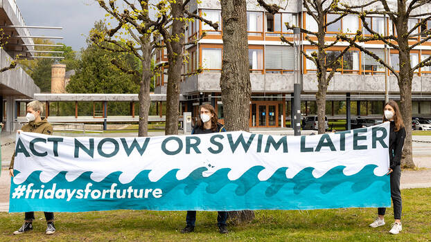 Aktivist*innen stehen auf der Wiese vor dem Gerichtsgebäude in Karlsruhe und haten ein Transparent mit der Beschriftung "Act Now Or Swim Later. #fridaysforfuture"